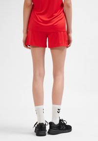 Röda sportshorts med en slät textur, kombinerade med svarta sneakers och vita strumpor med svart zickzackmönster.
