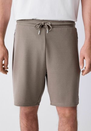Homme portant un short taupe à cordon et une chemise blanche, visible de la taille au milieu des cuisses sur un fond uni.