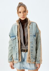 Jeansjacke mit beigem Kunstfellfutter, ausgestattet mit einer Knopfblende, zwei Brusttaschen und einer lockeren Passform, getragen über einem karierten Hemd.