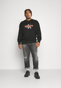 Hanorac negru cu un logo proeminent, combinat cu blugi gri uzati și pantofi sport negri. Aspectul este casual și modern.