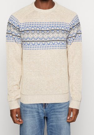 Beige strikket sweater med blå, grå og hvide geometriske mønstre over brystet og ærmerne, båret med blå jeans.