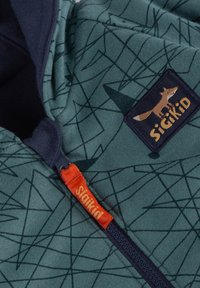 Felpa verde con zip caratterizzata da un motivo geometrico, dettagli in blu navy e una patch con una volpe. Il tirazip è di un luminoso arancione.