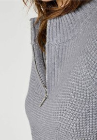 Pull gris à fermeture éclair avec texture côtelée et col montant. Comprend une fermeture éclair métallique argentée allant du cou à la poitrine.