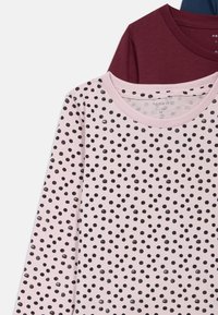 Camisa de manga longa em algodão rosa claro com pintas pretas. Apresenta um decote redondo canelado e está sobreposta a camisas burgundy e azul-marinho.