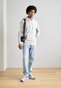 Lichtgrijze hoodie met een voorzak, gecombineerd met lichtgewassen jeans en zilveren sportschoenen. Het model heeft een zwarte crossbody tas bij zich.