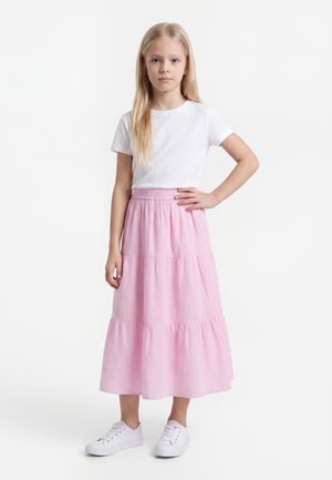 Νεαρό κορίτσι με μακριά ξανθά μαλλιά που φοράει λευκό μπλουζάκι, ροζ φούστα με στρώσεις και λευκά αθλητικά, στέκεται με το ένα χέρι στη μέση.