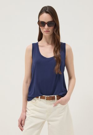 Vrouw met een schildpadkleurige zonnebril, een navyblauw mouwloos topje, crèmekleurige broek en een bruine riem, staand tegen een lichte achtergrond.