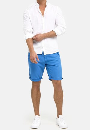 Hombre vestido con una camisa blanca de manga larga y botones, con las mangas arremangadas, pantalones cortos azules brillantes enrollados en el dobladillo y zapatillas blancas, de pie sobre un fondo blanco.