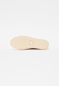 Sole of a sneaker in beige rubber with a textured grip pattern, featuring a small logo in the center. White background adds contrast.
