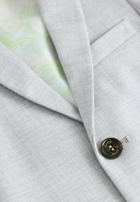 Blazer grigio chiaro con tessuto texture, rever a punte e un bottone scuro con dettagli incisi. La fodera interna presenta un motivo verde.