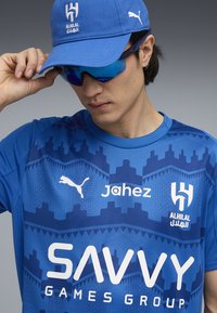 Blå atletisk trøje med geometriske mønstre, Puma-logo og sponsortekst. En matchende blå kasket og solbriller fuldender udseendet.