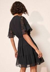 Juoda suknelė su permatomomis trumpomis rankovėmis, puoštomis nėrinių detalėmis, sluoksniuota suknele ir prigludusia liemene. Lygus, lengvas audinys su elegantiška, laisvai krentančia forma.