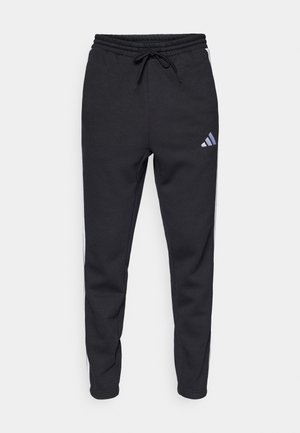 Pantalons de jogging noirs en tissu doux, avec une taille élastique, un cordon de serrage, et des bandes blanches sur les côtés avec un logo Adidas sur la cuisse.
