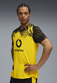 Joven con pelo trenzado posa con las manos en las caderas, vistiendo una camiseta de fútbol amarilla y negra del Borussia Dortmund con el logo de Vodafone.