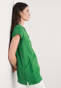 Tunic in lino verde con maniche corte, frontale bottonato, spacchi laterali e vestibilità comoda, abbinata a pantaloni bianchi e orecchini verdi.