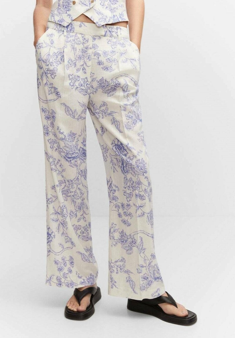 Femme portant un pantalon crème à jambes larges avec imprimé floral bleu et des sandales plateformes noires, mains dans les poches, debout sur un sol blanc.