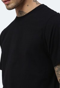 Schwarzes T-Shirt mit kurzen Ärmeln und rundem Ausschnitt, mit glatter Textur, das sichtbare Tattoos am Hals und am Arm aufweist.