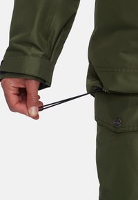 Giacca verde impermeabile con finitura testurizzata, con una mano che regola un cordino nero in vita per la regolazione della vestibilità.