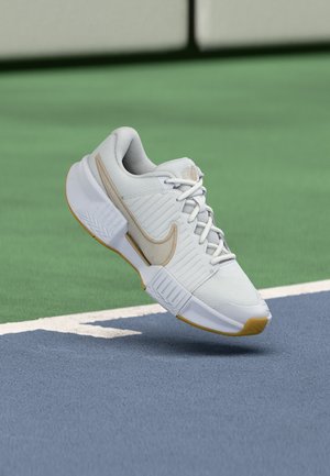 Weißer und beiger Nike-Sneaker, der über einem blau-grünen Tennisplatz mit weißen Begrenzungslinien schwebt.