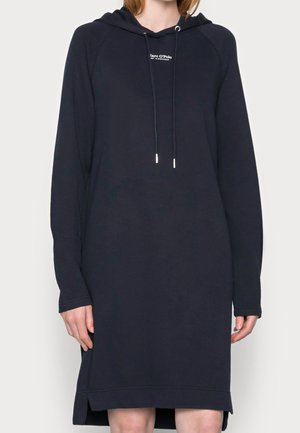 Femme portant une robe à capuche bleu marine à manches longues avec cordons et petit logo blanc sur la poitrine.