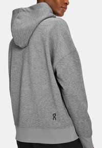Grauer Hoodie mit strukturierter Oberfläche, ausgestattet mit einer Kordelzugkapuze, gerippten Bündchen und einem Emblem auf der unteren linken Seite.