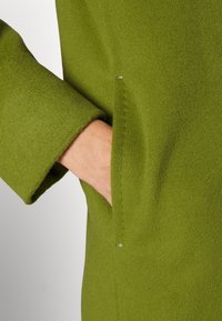 Grüner Wollmantel mit weicher Textur, ausgestattet mit einem umgeschlagenen Ärmelbündchen, einer seitlichen Tasche und einem dezenten Knopfdetail.