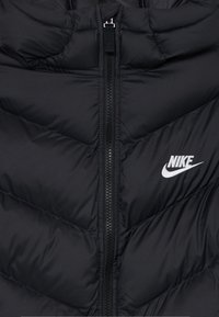 Juodas izoliuotas pūkinis švarkas su užtrauktuku priekyje, su chevron siuvinėjimu ir baltu Nike logotipu kairėje krūtinės pusėje.