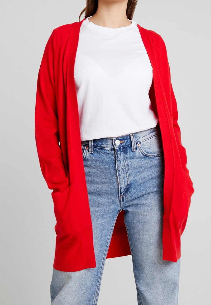 Femme portant un t-shirt blanc, un jean bleu taille haute et un long cardigan rouge ouvert devant avec des poches.