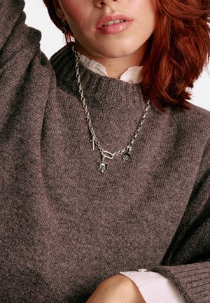 Grauer Strickpullover mit einem gerippten Halsausschnitt, getragen über einem weißen Hemd mit Kragen. Die silberne Kettencollier zeigt Anhänger in Form einer Fledermaus und anderer kleiner Formen.