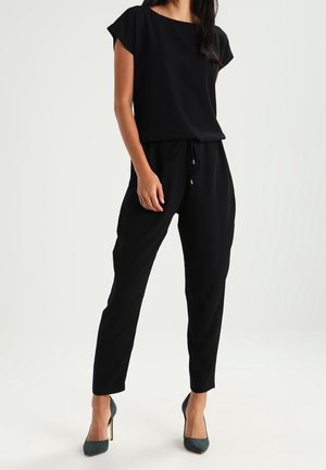 Vrouw draagt een zwart shirt met korte mouwen en zwarte broek met trekkoord, gecombineerd met donkergroene pumps, staand tegen een witte achtergrond.