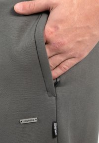 Gray athletic pants with a smooth texture, featuring a side pocket with a zipper. Includes a metal tag and a fabric label on the side.