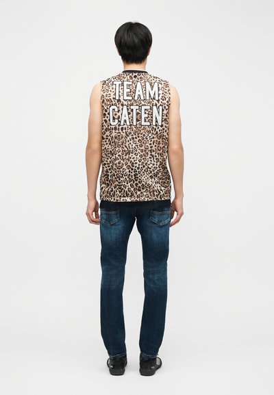 Personne debout vue de dos, portant un haut sans manches à imprimé léopard avec le texte "TEAM CATEN" et un jean bleu avec des chaussures noires.