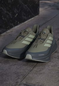 Paar graue und beige Adidas-Laufschuhe mit drei Streifen und Schnürsenkeln auf Steinboden in der Nähe einer dunklen Wand.