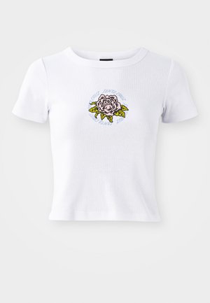 T-shirt branca de manga curta com design ajustado, apresentando um gráfico de rosa bordado centrado e texto circular em volta da flor.