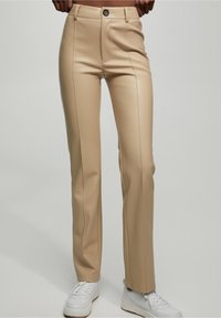 Pantalon beige sur mesure à coupe ajustée, avec plis devant et fermeture boutonnée, assorti à des baskets blanches. Texture lisse et lignes épurées.