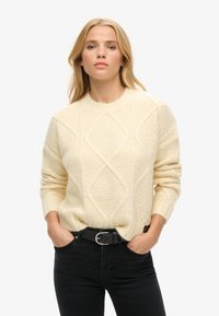 Kremowy sweter z fakturowanym wzorem w diamenty, okrągłym dekoltem i ściągaczami przy rękawach, zestawiony z czarnymi dżinsami i czarnym paskiem.