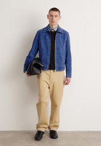 Μπλε σουέντ μπουφάν, μαύρο πουλόβερ, μπεζ παντελόνι. Κρατάει μια μαύρη τσάντα. Το μοντέλο στέκεται μπροστά σε απλή βαμμένη επιφάνεια. Καθημερινό, σύγχρονο ντύσιμο.