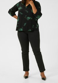 Sort bluse med grøn blomsterprint, v-hals og trekvartærmer; parret med stramme sorte jeans og spidse sorte hæle.