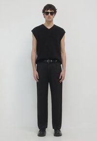 Schwarzer ärmelloser Pullover mit V-Ausschnitt, kombiniert mit locker sitzenden schwarzen Hosen und schwarzen Schuhen, ergänzt durch schwarze Sonnenbrillen.