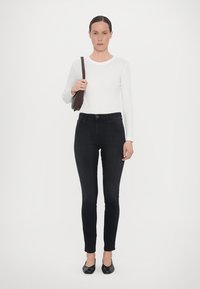Svarta slim fit-jeans med hög midja, kombinerat med en figurnära vit långärmad skjorta och svarta platta skor; brunt handväska över axeln.