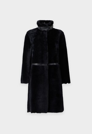 Femme portant un manteau en fourrure noir, un haut bleu marine, un pantalon beige à jambes larges et des ballerines noires à bout pointu, debout devant un fond clair uni.