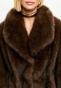 Brun faux fur-frakke med en bred, blød krave med diagonale mønstre, parret med et guld choker-halssmykke.