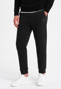 Schwarze Jogginghosen mit tailliertem Design, seitlichen Taschen und Kniefalten-Details. Kombiniert mit weißen Sneakern für einen Kontrast.