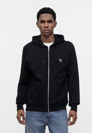 Jeune homme portant un sweat à capuche noir zippé avec un petit logo zèbre rayé en couleurs sur la poitrine et un jean bleu, debout sur un fond blanc uni.
