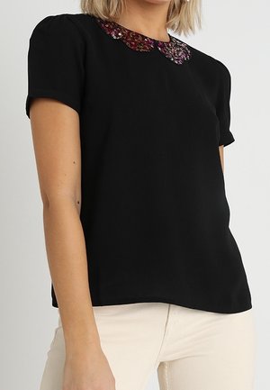 Femme portant un haut noir à manches courtes avec un col pailleté scintillant dans des nuances rouges et violettes, associé à un pantalon de couleur claire.