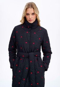 Prešívaný čierny kabát s vysokým golierom, červenými bodkami, s opaskom v páse a zácvakmi. Vzorovaná textúra s vlnitým prešívaním.