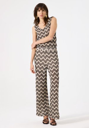 Ärmelloser Jumpsuit aus leichtem Stoff mit einem wellenförmigen schwarz-beigen Muster; weit geschnittene Beine; getragen mit schwarzen Riemchensandalen.