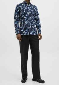 Camicia blu navy con bottoni e motivi floreali blu e bianchi, realizzata in tessuto liscio. Abbinata a pantaloni neri e scarpe nere.