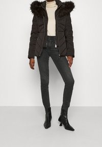 Sort pufferjakke med hætte med pelskant, lynlås foran og kviltet design, parret med mørkegrå højtaljede jeans og sorte ankelstøvler.