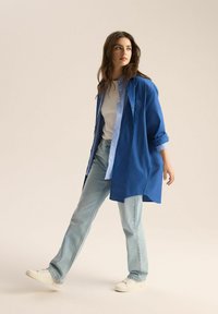 Niebieska, oversize koszula zapinana na guziki, nałożona na jasnoszary T-shirt, w połączeniu z jasnoniebieskimi dżinsami i białymi sneakersami. Gładki materiał, luźny krój.